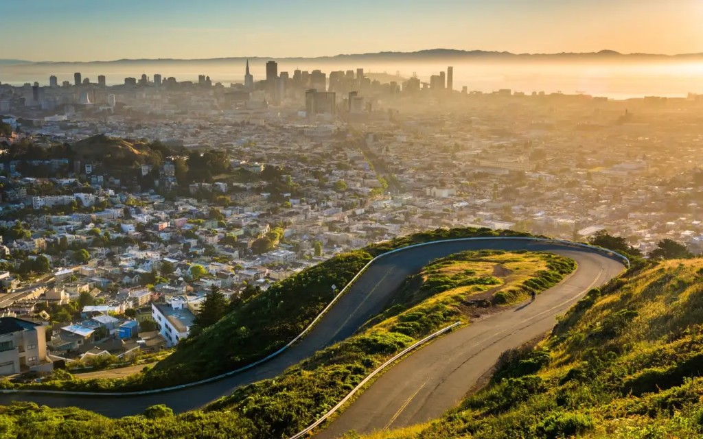 Panoramic golden-hour view from a winding hillside road over a city skyline, bay, and distant hills.