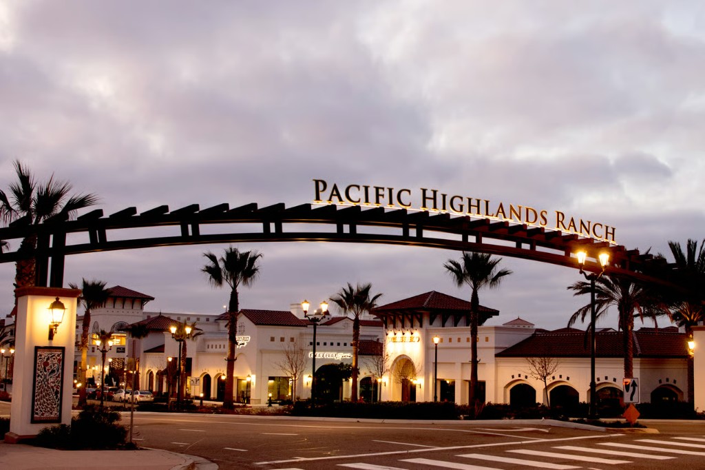 Pacific Highlands Ranch entrance at dusk: illuminated archway sign over the road, Mediterranean-style buildings, palm trees, and warm streetlights under a twilight sky.
