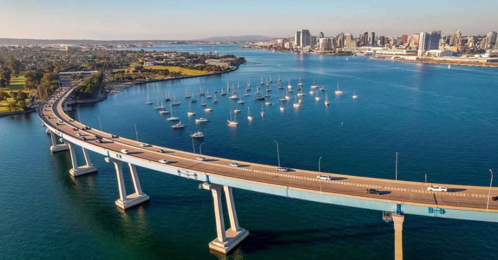 San Diego–Coronado Bridge over the bay at golden hour, with sailboats and the downtown San Diego skyline