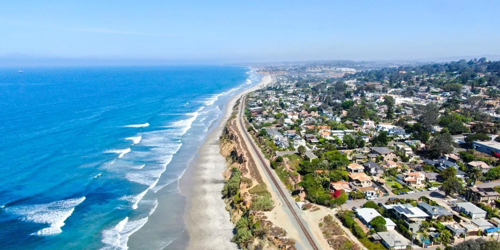 Aerial view of Del Mar coastline: Pacific Ocean, sandy beach, coastal bluffs, railroad along the shore, and residential neighborhood under a clear blue sky.