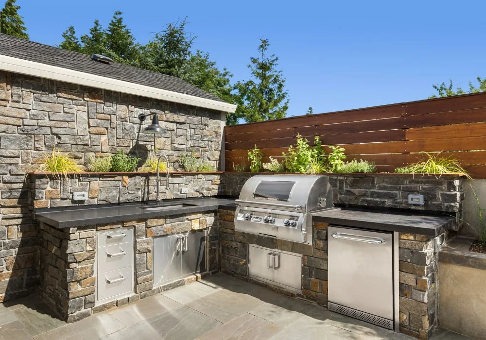 Luxury outdoor kitchen with stacked stone, dark countertops, built-in stainless grill, sink, and under-counter outdoor refrigerator on a patio.